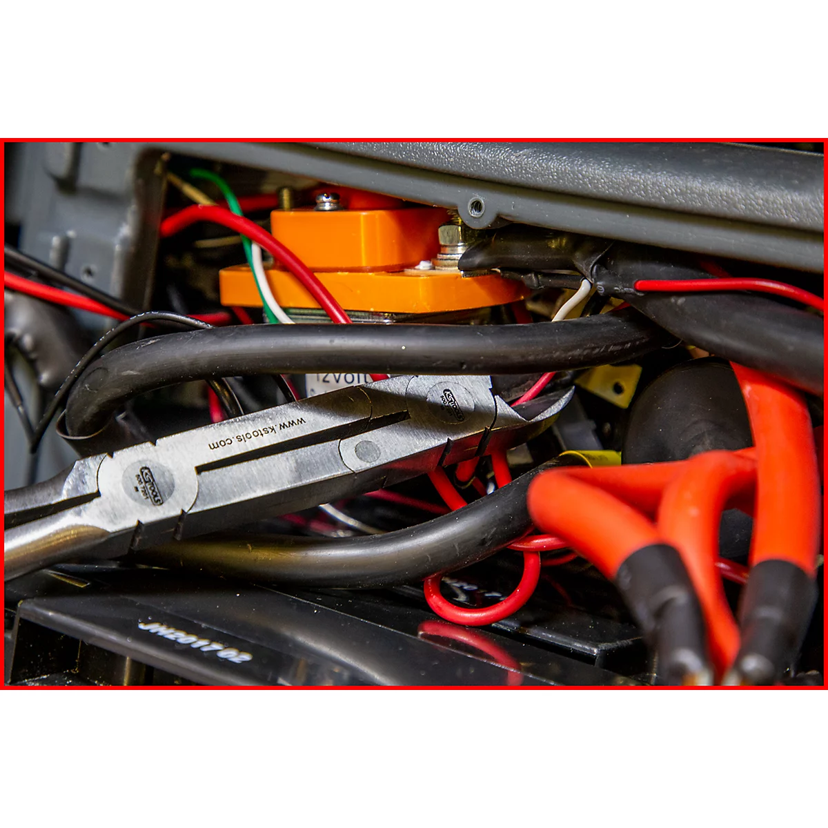 Nahaufnahme von Zangen, die an Kabeln in einem Motorraum arbeiten. Im Hintergrund orangefarbene elektrische Komponenten und rote Kabel.